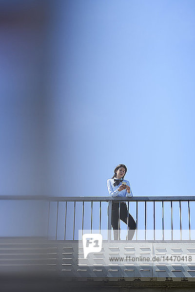 Geschäftsfrau steht auf Brücke vor blauem Himmel mit Smartphone