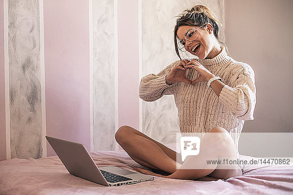 Young woman sitting on bed with laptop forming a heart with her hands during video chat