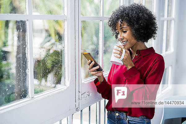 Lächelnde Frau mit Tasse Kaffee und Handy zu Hause am Fenster stehend