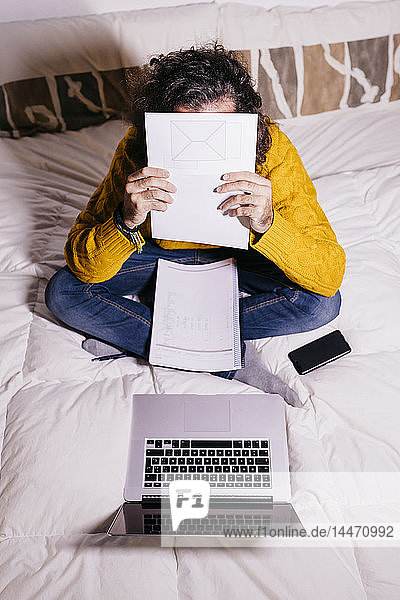 Young man working in bed with laptop covering his face with papers