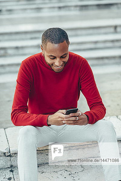 Smilig young man wearing red pullover sitting on stairs using cell phone