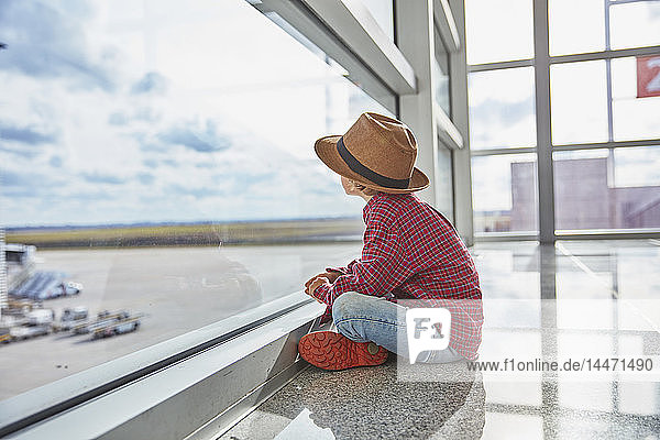 Junge sitzt auf dem Flughafen hinter einer Fensterscheibe und schaut auf den Flugplatz