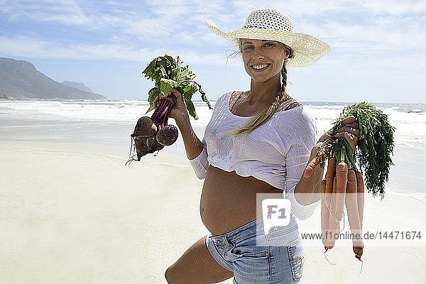 Portrait of smiling pregnant woman on the beach holding carrots and beetroot
