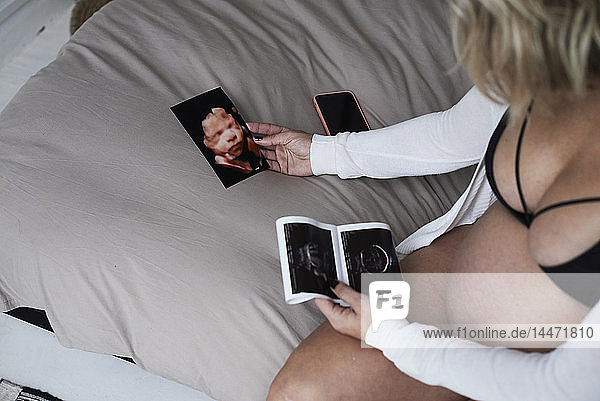 Schwangere Frau betrachtet Ultraschallbilder zu Hause im Sitzen auf dem Bett