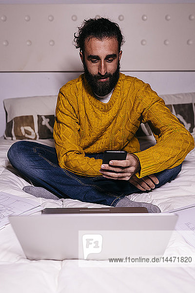 Young man working in bed at home with cell phone laptop and documents