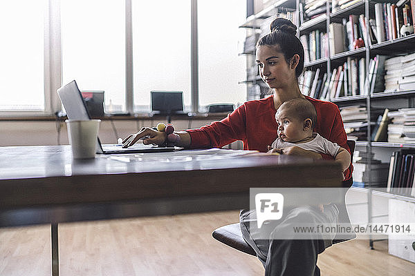 Berufstätige Mutter mit Baby auf dem Schoß  im Büro sitzend  mit Laptop