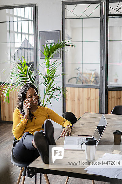Junge Geschäftsfrau sitzt mit Füßen auf dem Schreibtisch und telefoniert mit dem Handy