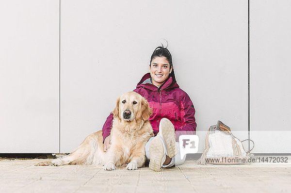 Porträt einer lächelnden jungen Frau mit ihrem Golden-Retriever-Hund an einer Wand sitzend