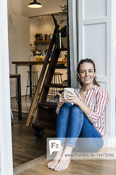 Porträt einer lächelnden Frau  die zu Hause mit einer Tasse Kaffee auf dem Boden sitzt