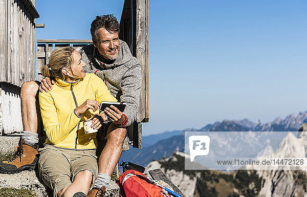 Hiking couple sitting in front of mountain hut  taking a break  looking at smartphone
