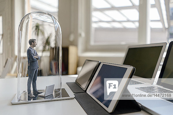 Geschäftsmann-Figur unter Glasglocke auf Schreibtisch stehend  mobilen Geräten zugewandt