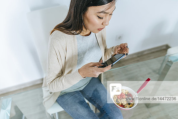 Junge Frau isst Müsli zum Frühstück  während sie ein Smartphone benutzt