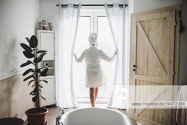 Frau im Bademantel mit Handtuch um den Kopf  die zu Hause am Fenster steht