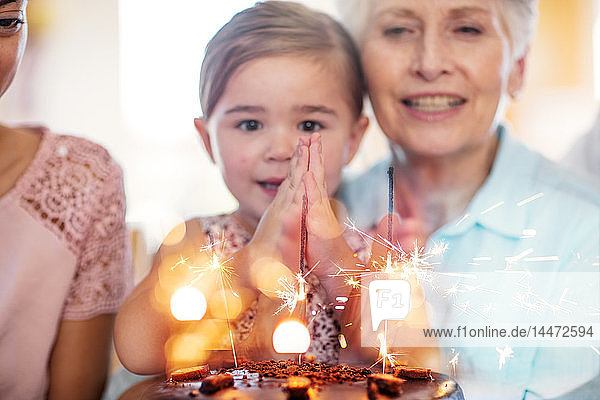 Kleines Mädchen schaut Wunderkerzen auf einem Geburtstagskuchen an und sitzt auf dem Schoß der Großmutter