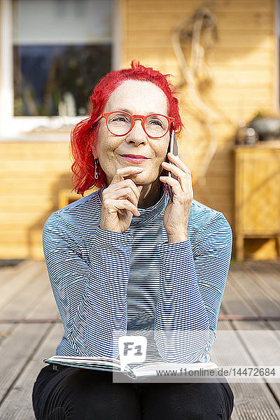 Porträt einer lächelnden älteren Frau mit rot gefärbten Haaren am Telefon  die auf der Terrasse vor ihrem Haus sitzt
