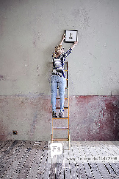 back view of woman on ladder hanging up picture frame in an unrenovated room of a loft