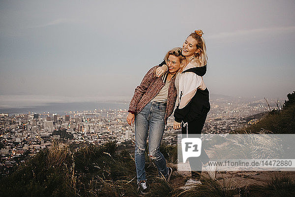 South Africa  Cape Town  Kloof Nek  two happy women embracing at sunset with cityscape in background