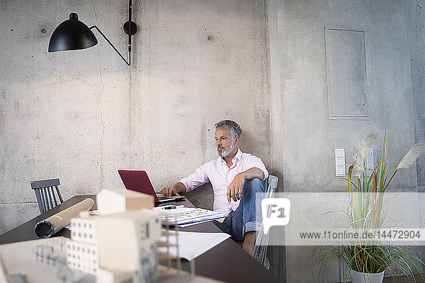 Businessman in a loft using laptop with documents and architectural model on table