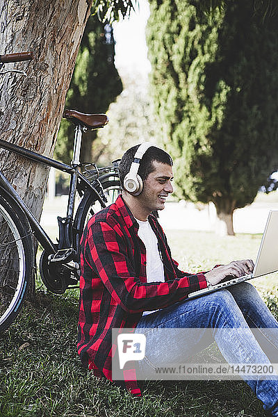 Junger Mann sitzt in einem Park  trägt Kopfhörer und benutzt Laptop neben dem Fahrrad