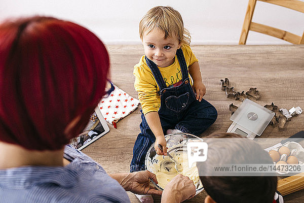 Lächelndes Kleinkindmädchen sitzt auf dem Küchentisch und bereitet zusammen mit Mutter und Bruder Teig
