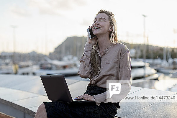 Happy young businesswoman using cell phone and laptop at the waterfront
