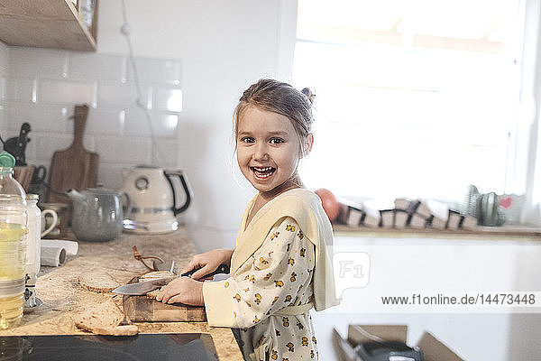 Porträt eines lachenden kleinen Mädchens  das in der Küche Brot schneidet