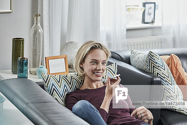 Lächelnde junge Frau benutzt Mobiltelefon zu Hause auf der Couch liegend