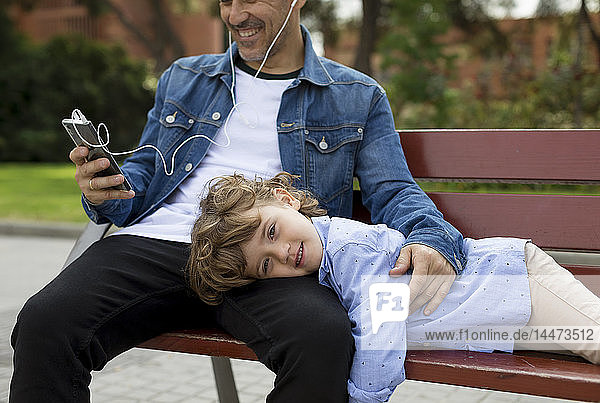 Porträt eines lächelnden Jungen mit Vater  der mit Handy und Ohrstöpseln auf einer Bank sitzt