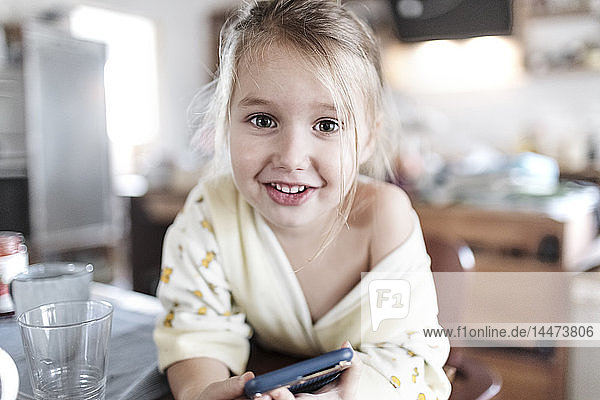 Porträt eines lächelnden kleinen Mädchens mit Smartphone in der Küche