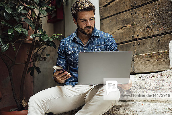 Geschäftsmann sitzt auf einer Treppe in einem Innenhof und benutzt Laptop und Mobiltelefon
