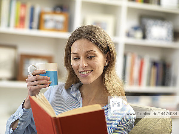 Lächelnde junge Frau mit einem heißen Getränk  die sich zu Hause beim Lesen eines Buches entspannt