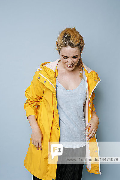 Lachende junge Frau in gelbem Regenmantel vor blauem Hintergrund