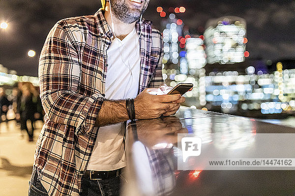 Grossbritannien  London  Nahaufnahme eines an einem Geländer lehnenden Mannes mit Mobiltelefon mit Stadtlichtern im Hintergrund
