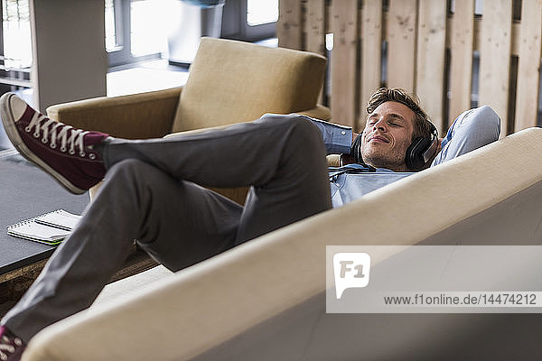 Geschäftsmann mit Kopfhörern auf der Couch liegend in Bürolounge
