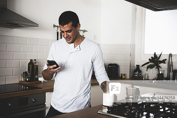 Smiling young man with cup of coffee using cell phone in kitchen at home