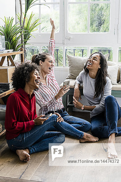 Drei unbekümmerte Frauen sitzen zu Hause mit Mobiltelefonen auf dem Boden
