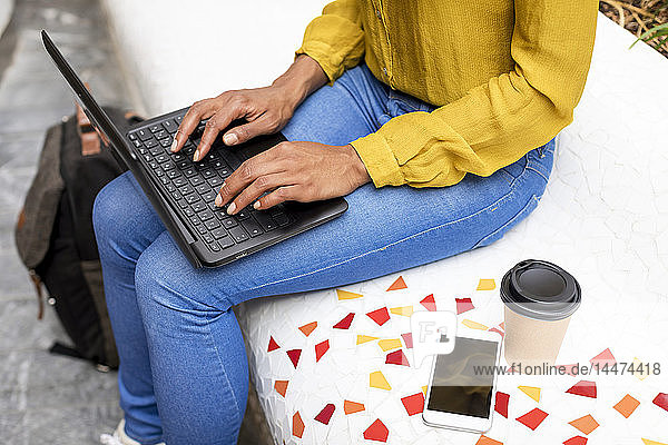 Frau sitzt auf Bank mit Smartphone und Kaffee zum Mitnehmen mit Laptop  Teilansicht