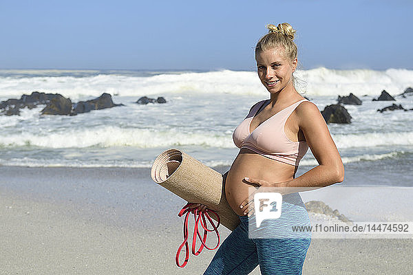 Porträt einer aktiven schwangeren Frau  die am Strand eine Gymnastikmatte hält