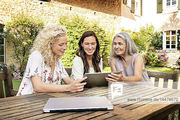 Drei Frauen unterschiedlichen Alters sitzen mit Tabletten am Gartentisch