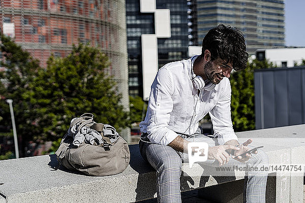 Lächelnder junger Mann sitzt auf einer Bank im Freien und benutzt sein Handy