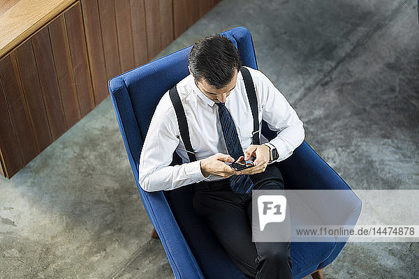 Top view of businessman sitting in blue armchair using smartphone
