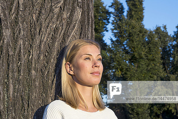 Porträt einer tagträumenden blonden Frau  die sich an einen Baumstamm lehnt und in die Ferne schaut