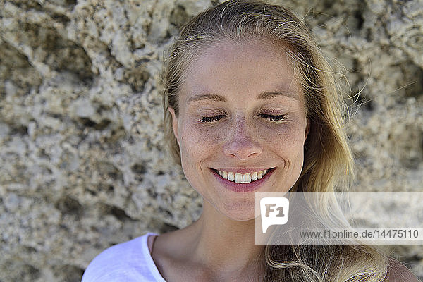 Bildnis einer lächelnden blonden Frau mit geschlossenen Augen vor felsigem Hintergrund