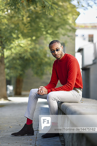 Portrait of fshionable young man wearing red pullover and sunglasses sitting on stone bench
