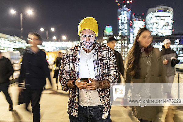 Großbritannien  London  lächelnder Mann  der nachts auf sein Telefon schaut  während in der Nähe verschwommene Menschen vorbeigehen