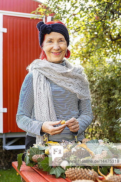Porträt einer lächelnden älteren Frau mit rot gefärbten Haaren beim Basteln an der Herbstdekoration im Garten
