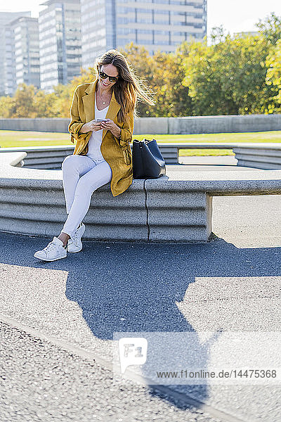 Lächelnde Frau mit Tasche sitzt auf einer Bank in der Stadt und benutzt ihr Handy