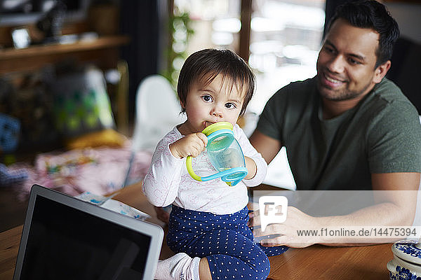 Portrait of baby girl sitting on table drinking water at home with father in background