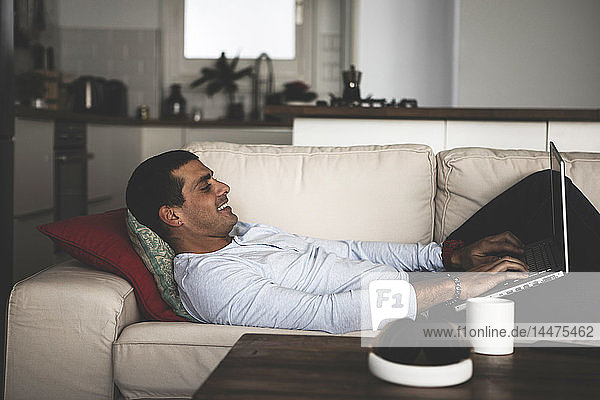 Smiling young man lying on sofa at home using laptop