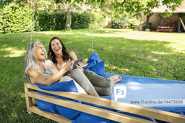 Zwei lachende Frauen entspannen sich mit Tabletten auf einem hängenden Bett im Garten